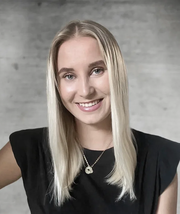 Smiling young woman with long straight blonde hair wearing a black sleeveless top and a gold necklace with a pendant standing against a gray textured background.