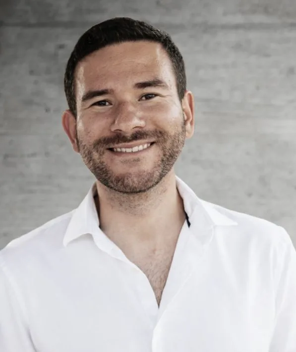 Smiling man with short dark hair and light beard wearing a white button-up shirt against a plain gray background